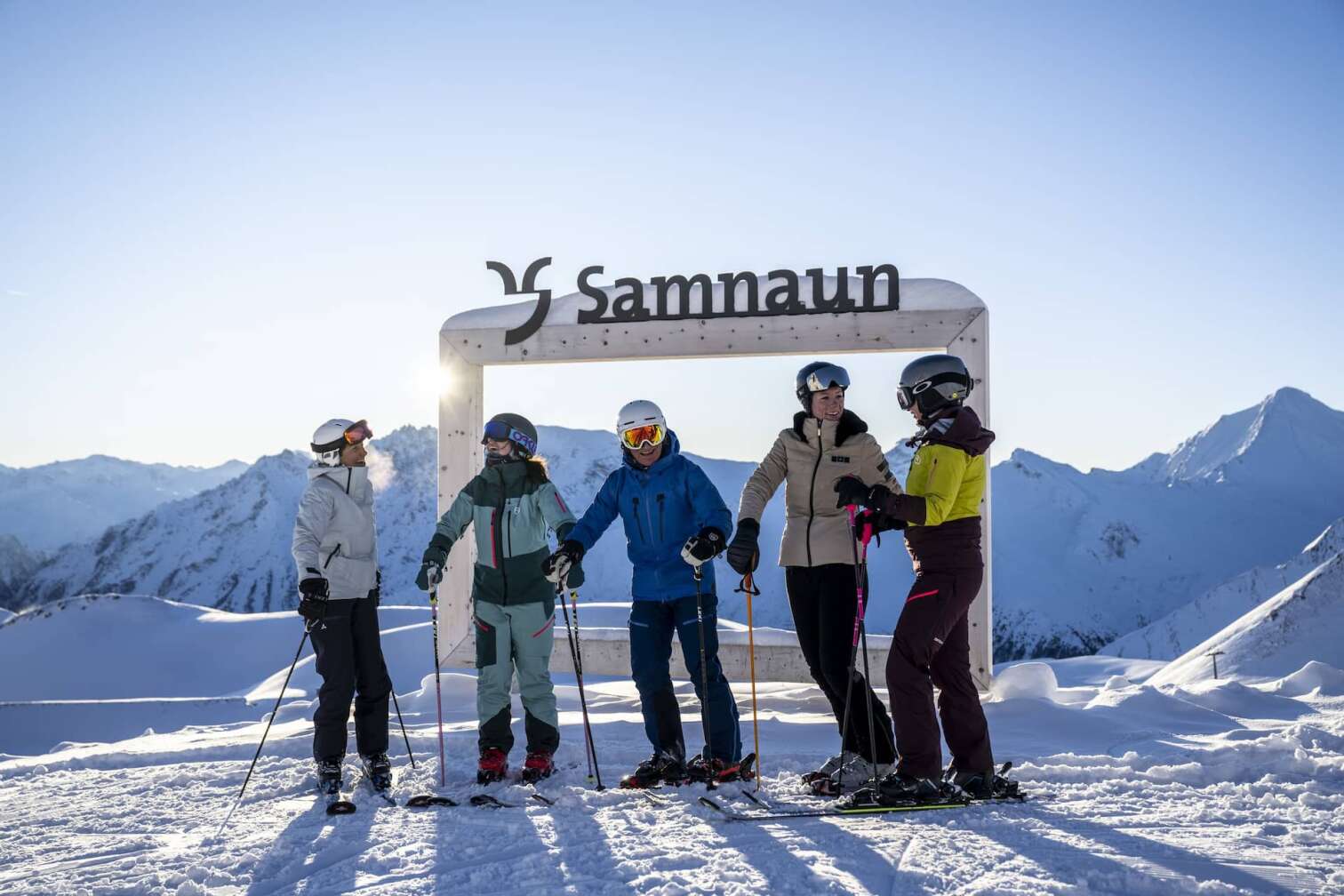 eine Gruppe von Skifahrern vor dem Schild "Samnaun" vor dem Bergpanorama der Silvretta Arena