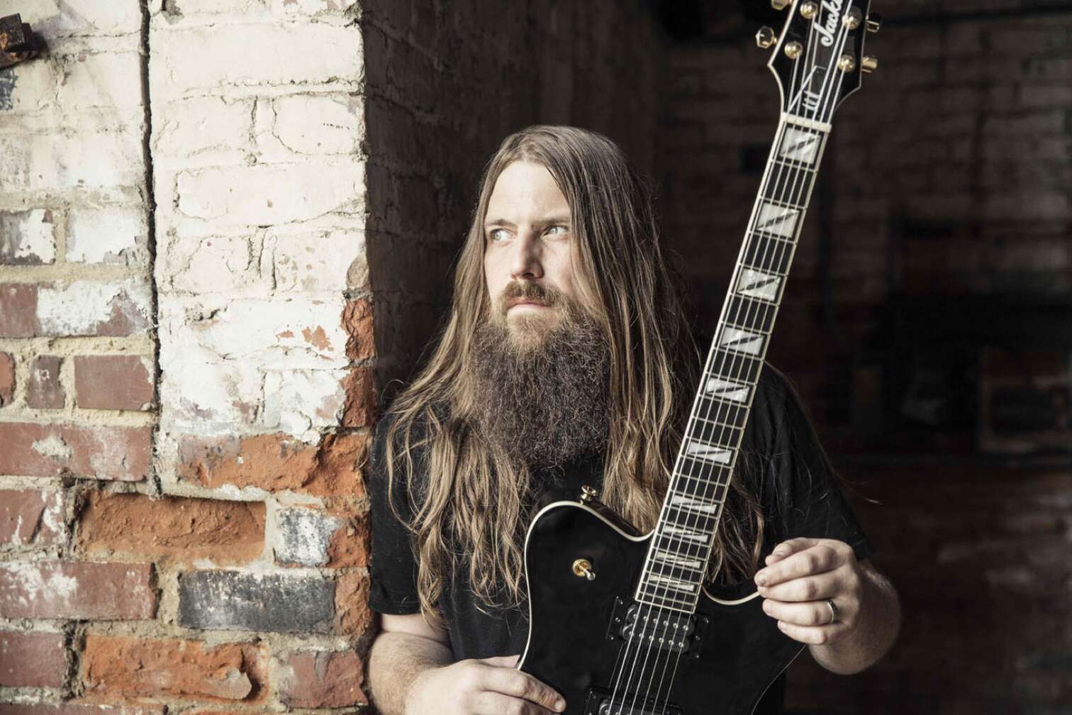 Mark Morton hält eine Gitarre und posiert vor einer Backsteinwand